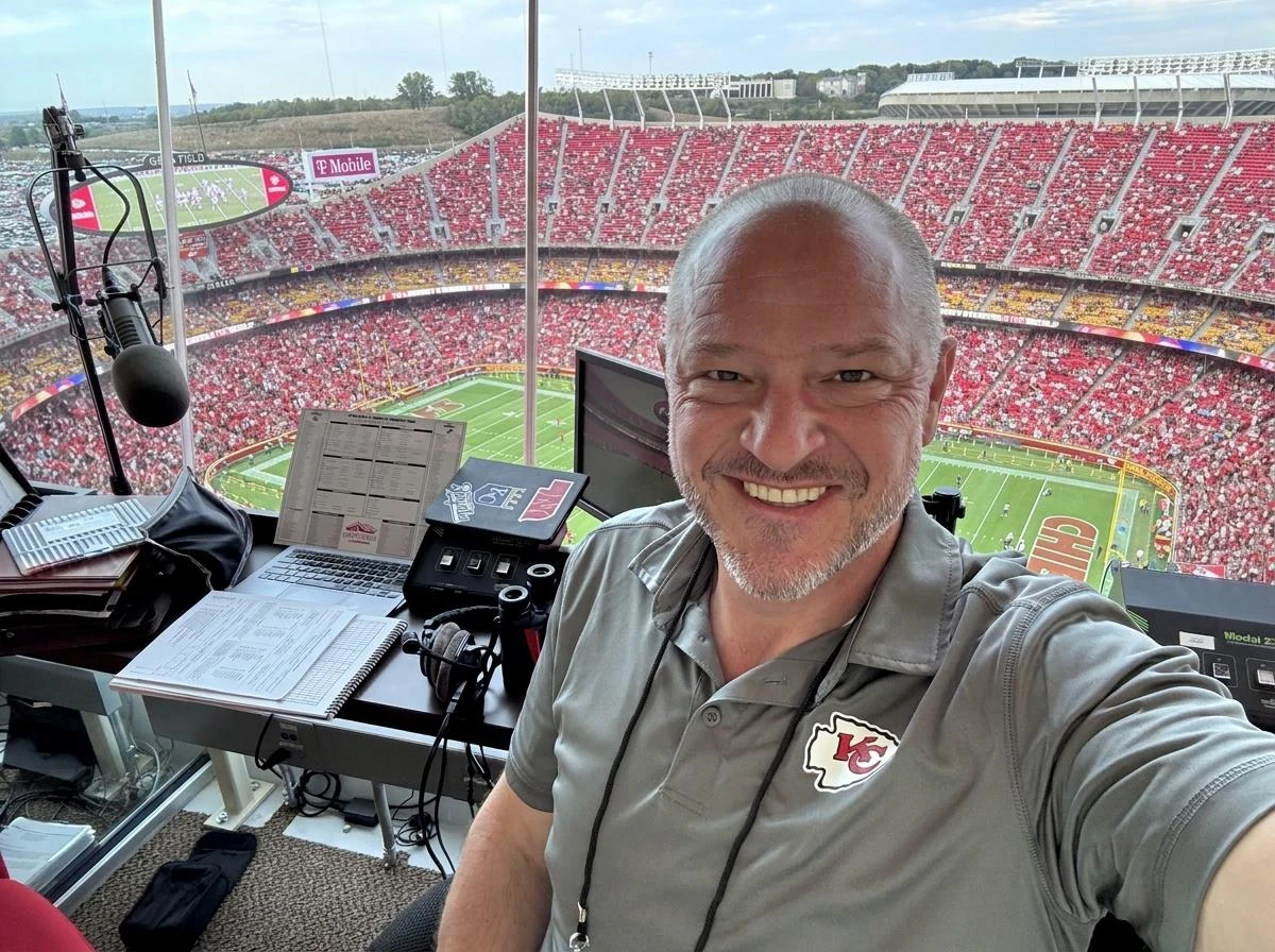 Chris Kamler smiling in a broadcast booth overlooking a packed football stadium